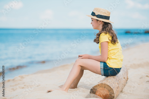 side view of cute little girl sitting on wooden trunk on sandy beach