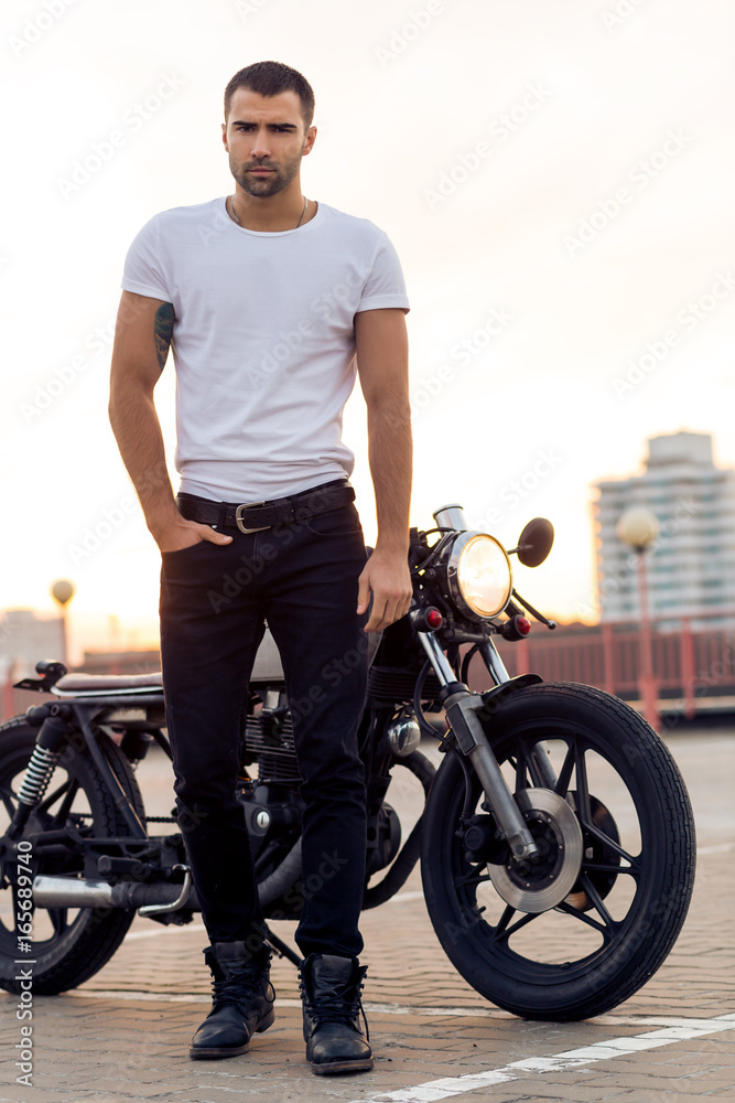 Fototapeta premium Close up of a handsome rider guy in white blank t-shirt look to camera near classic style cafe racer motorcycle at sunset. Bike custom made in vintage garage. Brutal urban lifestyle. Outdoor portrait.