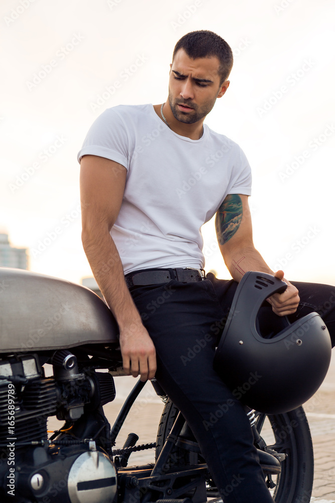Close up of a handsome rider man in white blank t-shirt look to the side near classic style cafe racer motorbike at sunset. Vintage bike custom. Brutal urban lifestyle. Outdoor portrait.