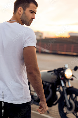 Fotótapéta Sporty biker handsome rider guy in white t-shirt want to ride his classic style cafe racer motorbike on rooftop at sunset