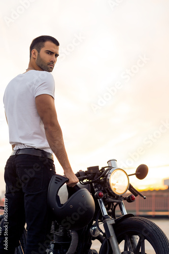 Sporty biker handsome rider man in white t-shirt want to ride his classic style cafe racer motorbike on rooftop at sunset. Vintage bike custom made in garage. Brutal urban lifestyle. Outdoor portrait.