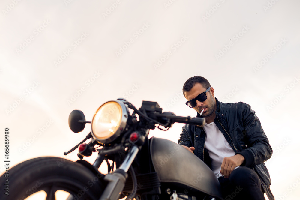 Handsome rider male with beard and mustache in black biker jacket, white shirt and fashion sunglasses smoke cigaret and sit on classic style cafe racer motorcycle at sunset. Brutal urban lifestyle. Handsome rider male with beard and mustache in black biker jacket, white shirt and fashion sunglasses smoke cigaret and sit on classic style cafe racer motorcycle at sunset. Brutal urban lifestyle.