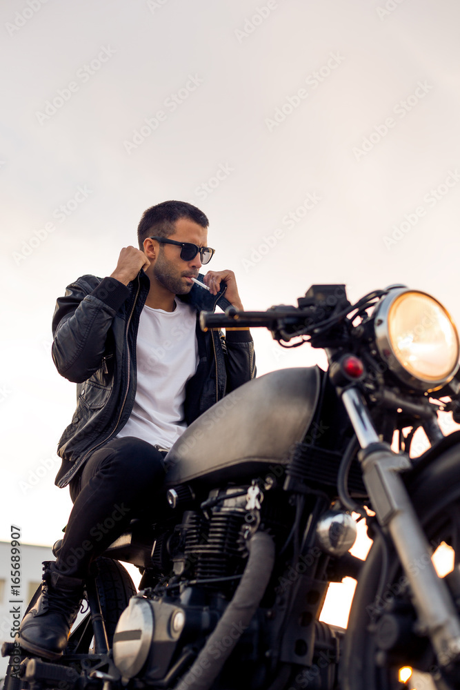 Handsome young rider man with beard and mustache in black fashion sunglasses smoking cigaret and correct biker jacket sit on classic style cafe racer motorbike at sunset. Brutal fun urban lifestyle. Handsome young rider man with beard and mustache in black fashion sunglasses smoking cigaret and correct biker jacket sit on classic style cafe racer motorbike at sunset. Brutal fun urban lifestyle.