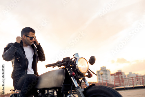 Handsome rider guy with beard and mustache smoking cigaret and correct biker jacket sit on classic style cafe racer motorbike at sunset. Bike custom made in vintage garage. Brutal fun urban lifestyle.