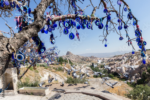 Evil Eye Beads on Tree and Fairy tale chimneys on background of blue sky in Guvercinlik Valley,Goreme,Turkey.Branches of the old tree decorated with the eye-shaped amulets photo