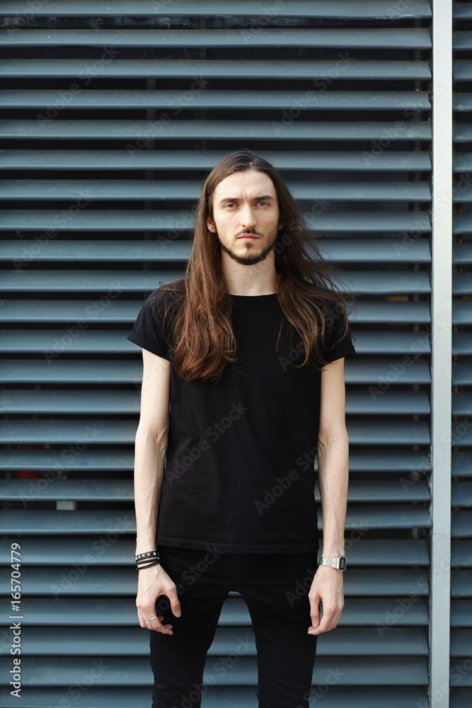 Style and fashion concept. Cropped outdoor portrait of handsome European  male model with beard and long hairstyle, dressed in black t-shirt and  skinny jeans, looking at camera with serious expression Stock Photo |