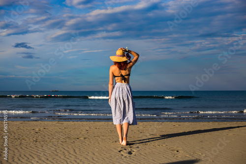 Young woman good looking redhead girl posing and turning back to the camera on beach barefoot with arm holding the hat, bikini and hat with blue sky and dreamy sea waves in background