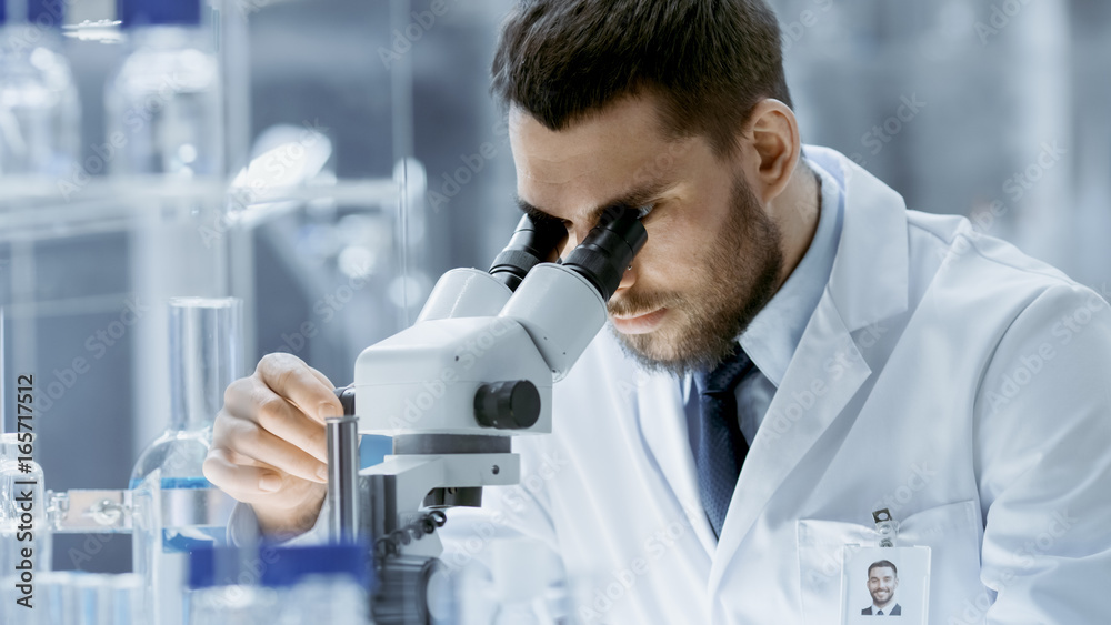 Close-up Shot of Research Scientist Adjusts His Microscope. He's Working in a High-End Modern Laboratory with Beakers, Glassware, Microscope and Working Monitors Surround Him.