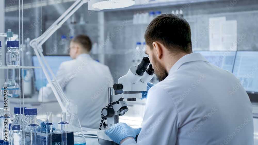 Shot of Research Scientist Adjusts His Microscope. He's Working in a High-End Modern Laboratory with Beakers, Glassware, Microscope and Working Monitors Surround Him. Shot of Research Scientist Adjusts His Microscope. He's Working in a High-End Modern Laboratory with Beakers, Glassware, Microscope and Working Monitors Surround Him.