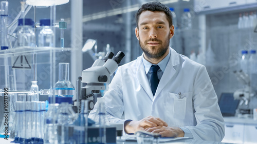 Young Male Research Looks into Camera and Smiles. He's Sitting in a High-End Modern Laboratory with Beakers, Glassware, Microscope and Working Monitors Surround Him.