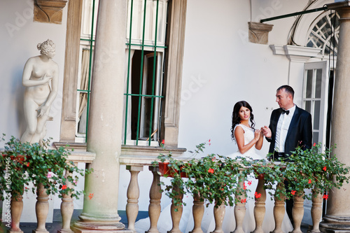 Amazing young attractive newly married couple walking and posing in the downtown with beautiful architecture and flowers on the background on their wedding day.