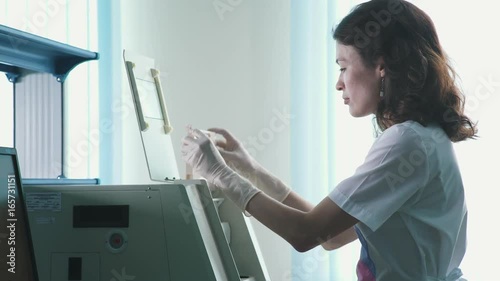 A woman in sterile clothes (white coat and gloves) works in a scientific laboratory and stands near the equipment, adjusts it. Work in scientific production: laboratory assistant in the process photo