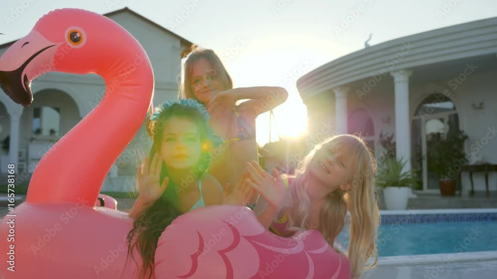 small girlfriends on summer vacation, sweet girls lie on inflatable pink flamingo near pool, spoiled rich childs in backlight outdoors, children have fun in background villa, kids celebritie