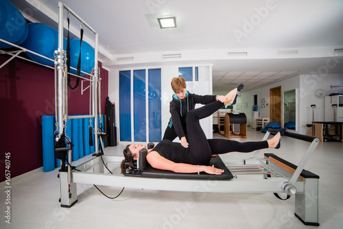 Young woman exercising on pilates device reformer with instructor at gym