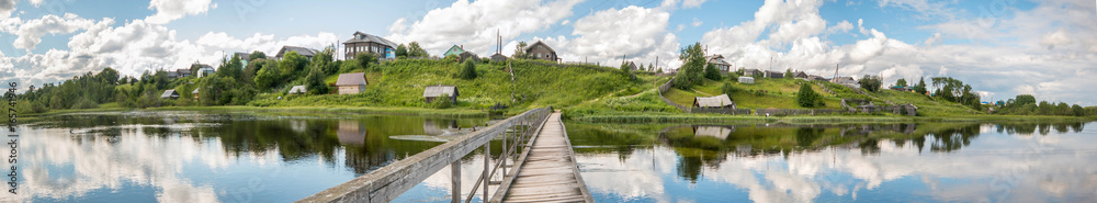 north Russian village Isady. Summer day, Emca river, old cottages on the shore, old wooden bridge and clouds reflections.