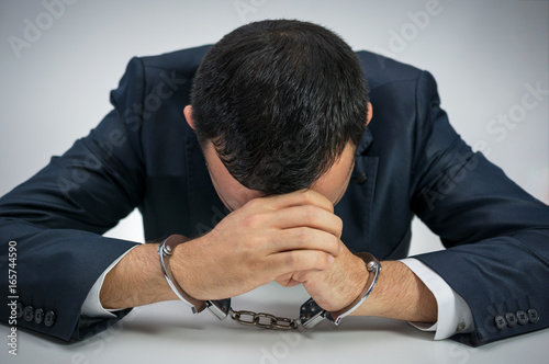 White young businessman handcuffed. He is sitting at a table with face between his palms showing guilt and frustration