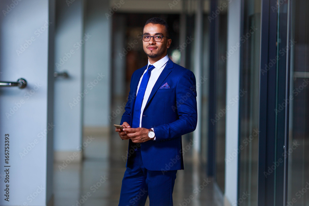 Young confident business man in suit and eyeglasses standing at the office with smartphone in his hand Young confident business man in suit and eyeglasses standing at the office with smartphone in his hand