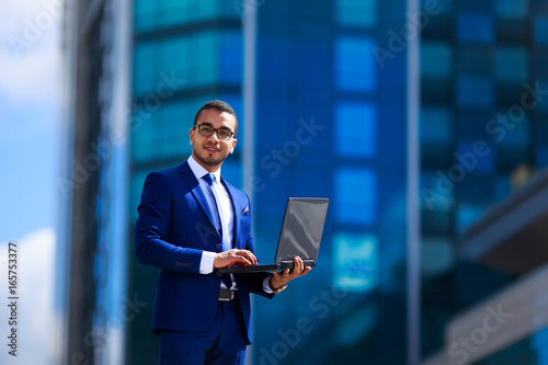 Low angle view of young confident businessman holding laptop and expressing positivity while standing outdoors with office building in the background