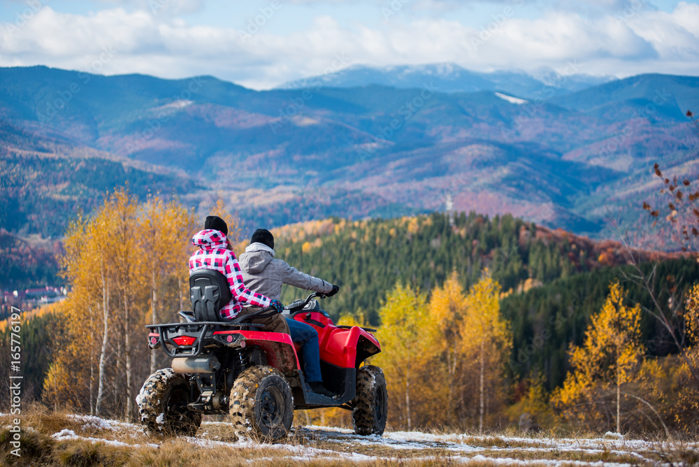 Fototapeta premium Rear view of man and woman in winter clothing riding a quad bike atv at the hill on the background of beautiful landscape mountains and forests.