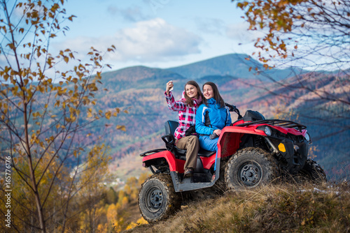 Smiling women in jackets on red ATV at the hill makes selfie on the phone with mountains in blurred background