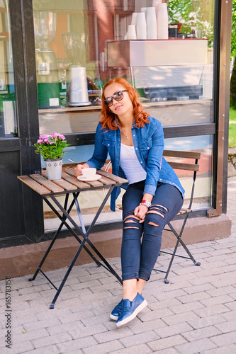 Redhead female drinks coffee at the table in a cafe on a street.