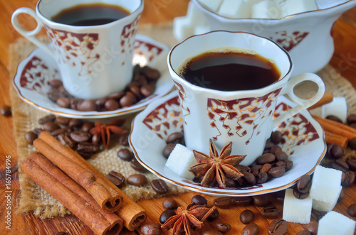 A two cups of black coffee with sugar bowl among the sackcloth, cinnamon bark, whole grains of coffee, anise stars and lump sugar on wooden table