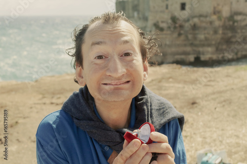 An ugly man kneels in front of her girlfriend at a romantic place and shows an engagement ring. Point-of-view of the woman. Funny comedy scene.
 photo