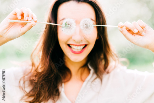 Portrait of young happy cheerful smiling and laughing beautiful brunette cute girl with long hair enjoying summer on vacation in park. Female hiding eyes with two dandelions. Guess who. Invisible life photo