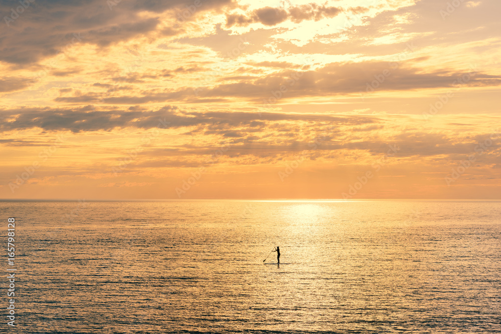 The concept of loneliness. Silhouette of a woman far away on the horizon, floating on a surfboard with a paddle in her hands, on the background golden clouds. The concept of loneliness. Silhouette of a woman far away on the horizon, floating on a surfboard with a paddle in her hands, on the background golden clouds.