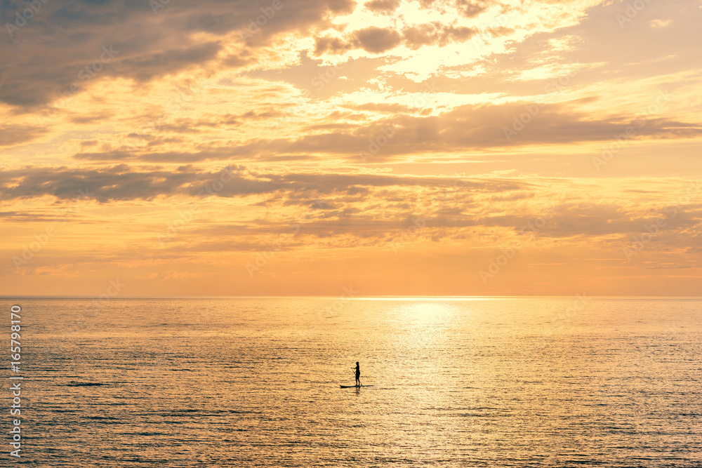 The concept of loneliness. Silhouette of a woman far away on the horizon, floating on a surfboard with a paddle in her hands, on the background golden clouds.