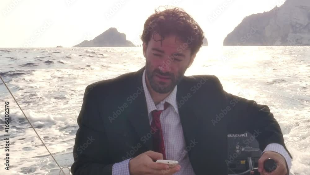 Man in suit with a briefcase texting on his smartphone while driving a boat in the middle of the ocean