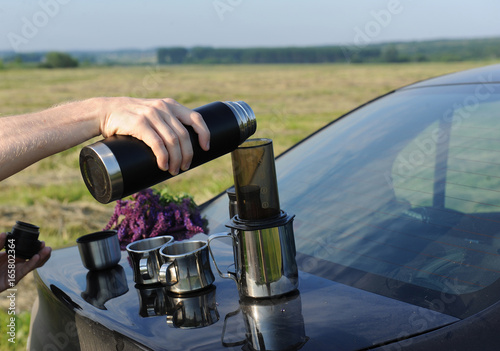 Items for alternative brewing of coffee in travel on the trunk of a black car. Summer landscape in the field