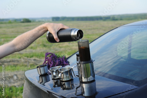 Items for alternative brewing of coffee in travel on the trunk of a black car. Summer landscape in the field
