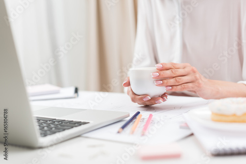cropped shot of businesswoman holding cup of coffee while working with laptop at workplace