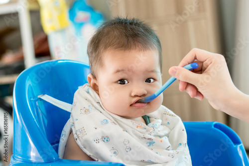 Portrait of happy asian mother feeding her cute little baby and looking in camera indoors