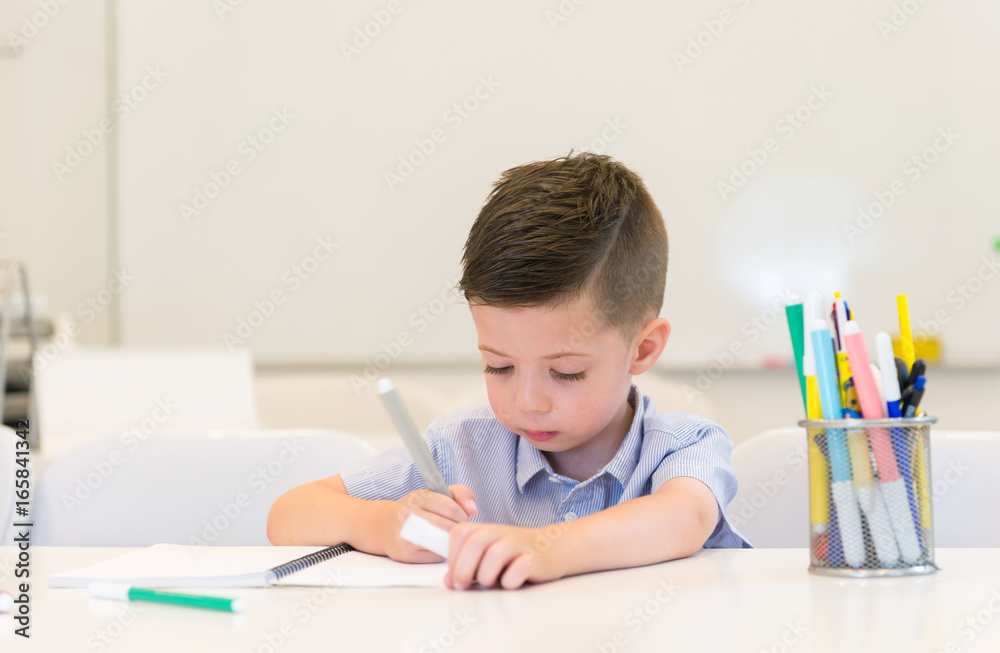 child preschooler boy drawing on booklet ,while sitting on the desk in the classroom child preschooler boy drawing on booklet ,while sitting on the desk in the classroom