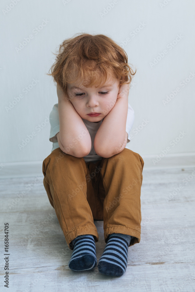 A little cute red-haired sad boy in a white T-shirt was offended and upset, sitting on the floor