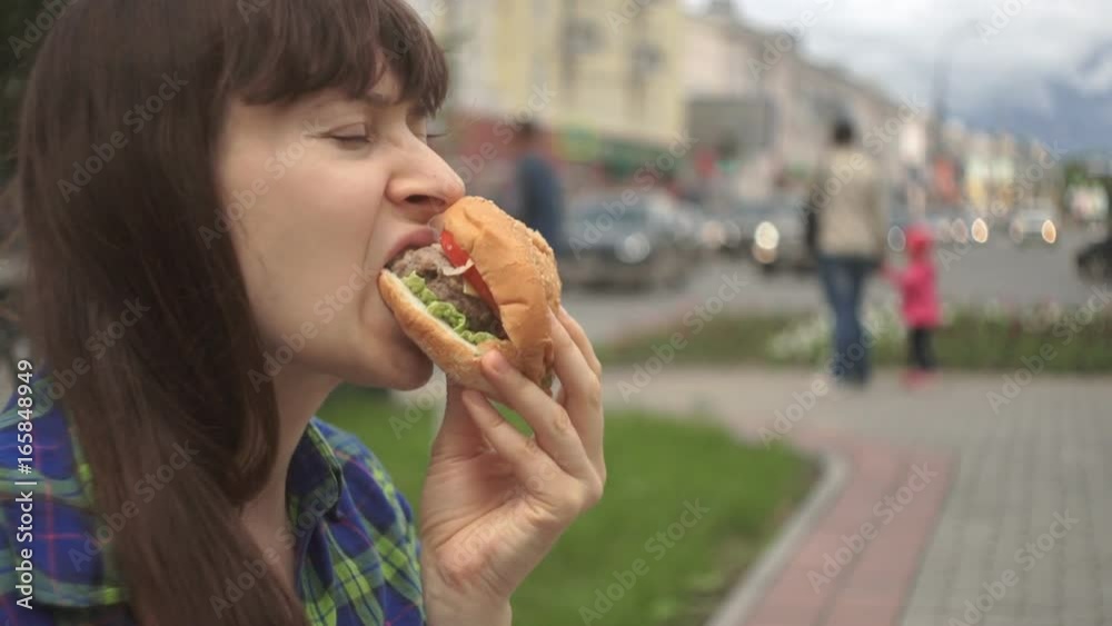 Close-up shot of a young attractive brunette woman in a plaid shirt eating a hamburger outside in the background of the busy traffic of people on the road and cars on the freeway, slow motion. Close-up shot of a young attractive brunette woman in a plaid shirt eating a hamburger outside in the background of the busy traffic of people on the road and cars on the freeway, slow motion.
