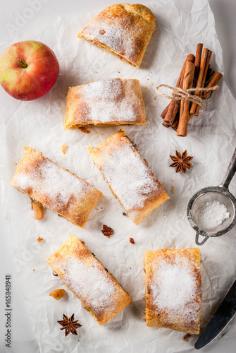 Home autumn, summer baking, puff pastries. Apple strudel with nuts, raisins, cinnamon and powdered sugar. On white marble table. Sliced, with ingredients. Copy space top view