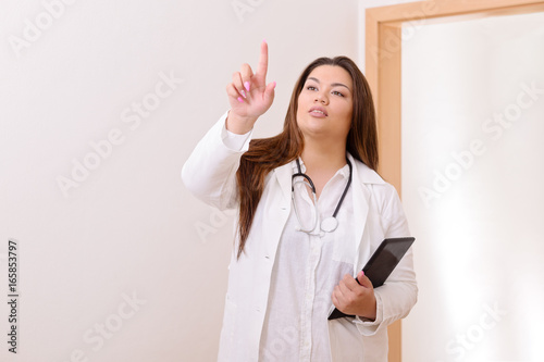 Young female hispanic doctor standing in a hospital hallway  holding digital tablet  pointing her finger in the air