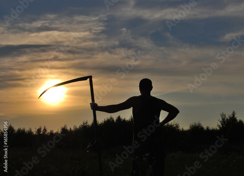 Silhouette of a man with a scythe against the sun in the evening in the field © all2014