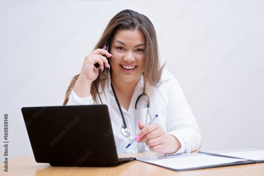 Young smiling female hispanic doctor sitting behind the desk talking on the mobile phone Young smiling female hispanic doctor sitting behind the desk talking on the mobile phone