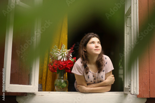 teenager girl look out of the window at morning from cottage summer house with rose bouquet in vase photo