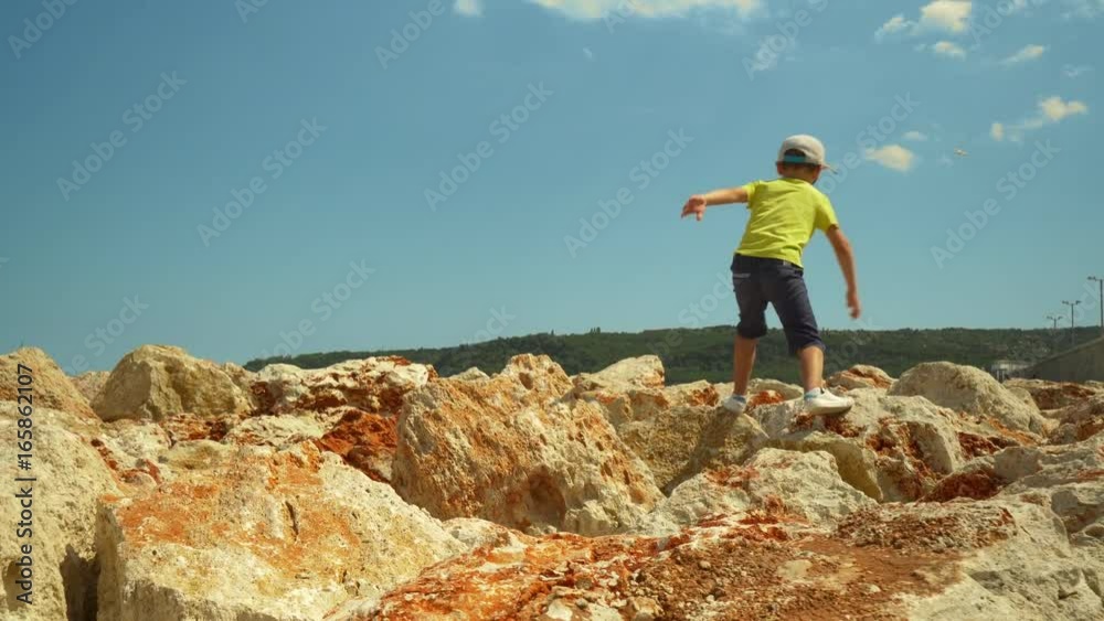 A happy boy walks along the stony seashore. A child jumping on a large yellow stone against a blue sky on the seashore
A happy boy walks along the stony seashore. A child jumping on a large yellow stone against a blue sky on the seashore