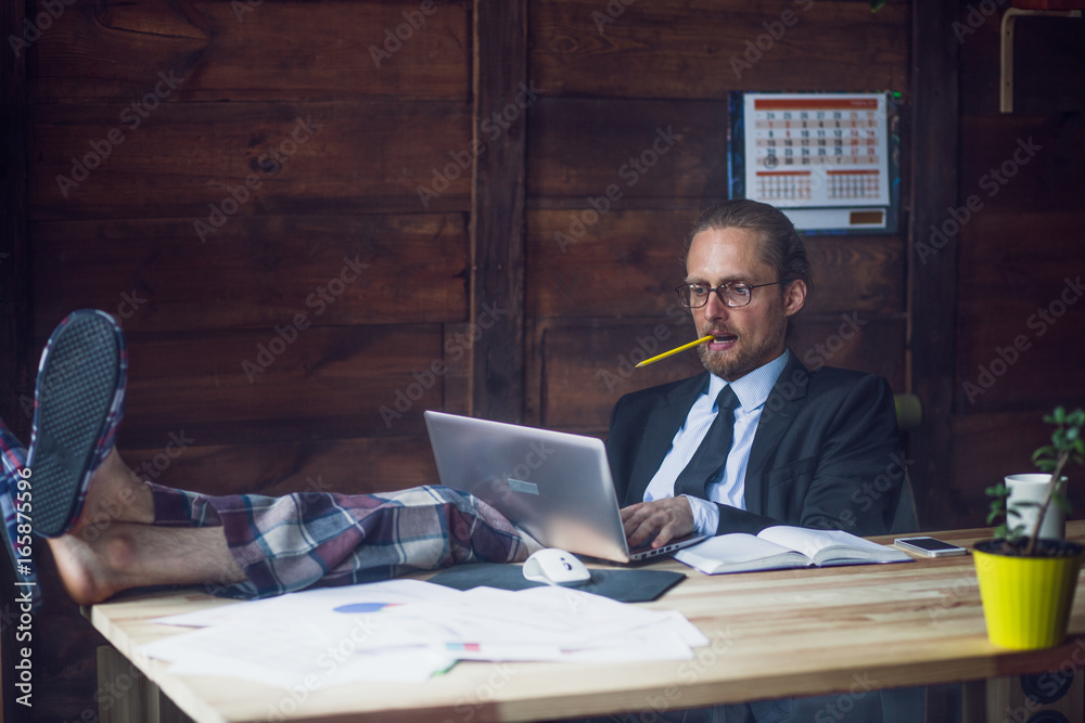 Freelance wearing business jacket, shirt and pajamas trousers. Man at home office holding legs on wooden desk while working with laptop. Toned image. - obrazy, fototapety, plakaty 
