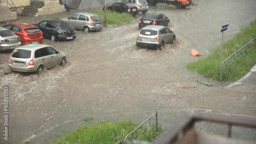 Cars drive along a flooded road in heavy rain, flood after heavy rainfall, streams of water come to the road from courtyards, slow motion.