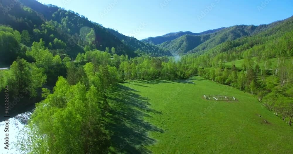 Aerial UHD 4K view. Mid-air flight over fresh mountain river and meadow at sunny summer morning. Green trees and sun rays on horizon. Rural dirt road below. Aerial UHD 4K view. Mid-air flight over fresh mountain river and meadow at sunny summer morning. Green trees and sun rays on horizon. Rural dirt road below.
