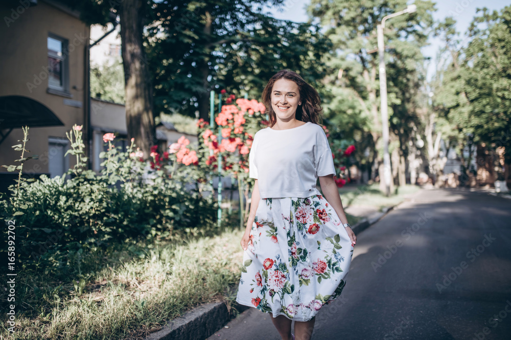 Beautiful sensual brunette young woman in white blouse and skirt with flowers walk on the street close to red roses bush. Big green trees on the background. Beautiful sensual brunette young woman in white blouse and skirt with flowers walk on the street close to red roses bush. Big green trees on the background.