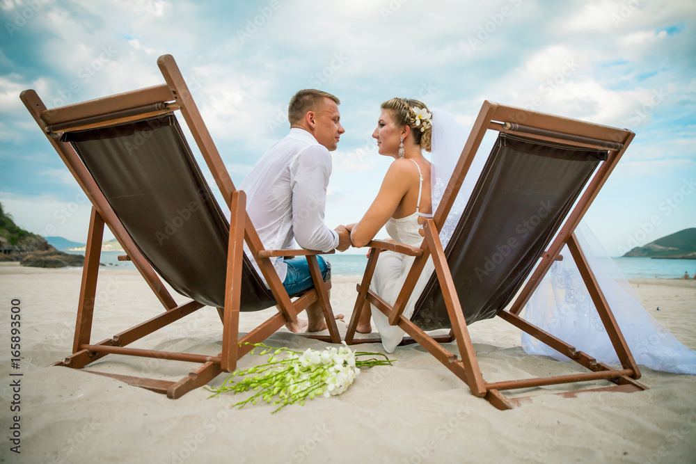 Bride and groom sitting in lounge chair by the sea on their wedding day