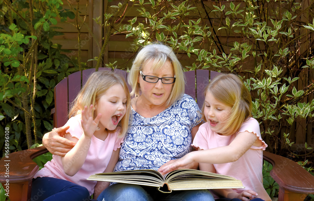 Twin sisters take time out for a story time with grandma. They sit together in a wooden lawn chair listening to a story told to them by their grandparent. Twin sisters take time out for a story time with grandma. They sit together in a wooden lawn chair listening to a story told to them by their grandparent.